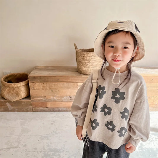 Child wearing a beige floral sweatshirt and hat in a minimalistic room with wooden boxes and baskets.